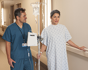 Woman patient walking in hospital hall with IV pole and nurse.