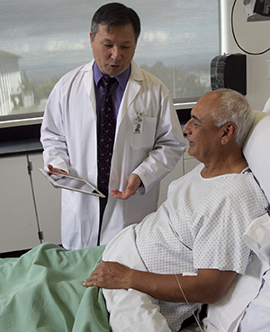 Health care provider with electronic tablet talking to man in hospital bed.